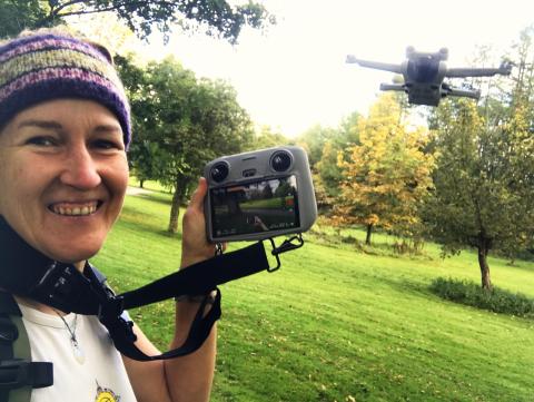 Smiling woman operating drone with remote control above grass and small trees